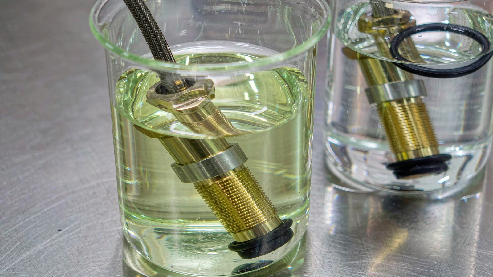Nickel-plated brass cable glands submerged in beakers of test liquid on a laboratory bench