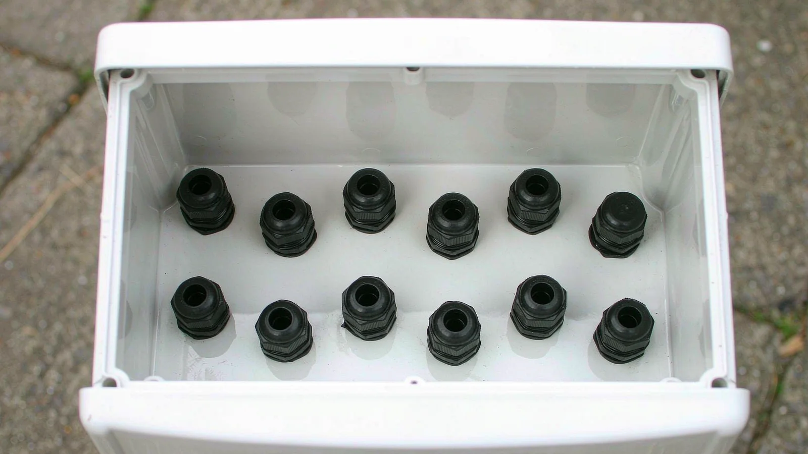 Two offset rows of black cable glands fitted into the bottom of a PV box, with blanking plugs closing the spare positions