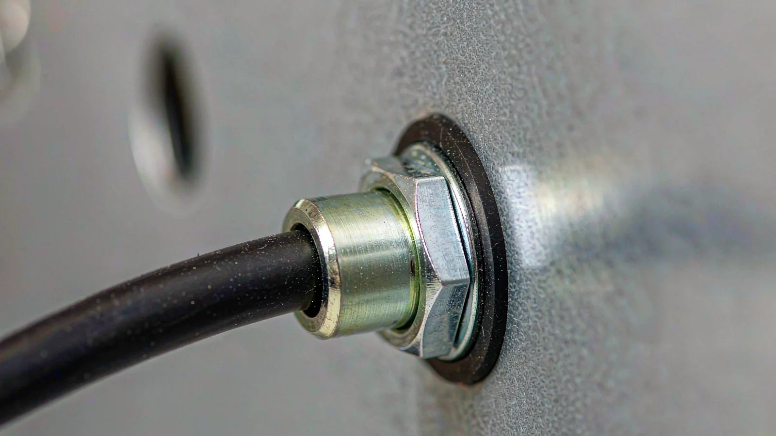 Nickel-plated brass cable gland passing through a sheet-steel enclosure wall with its black sealing washer compressed against the face