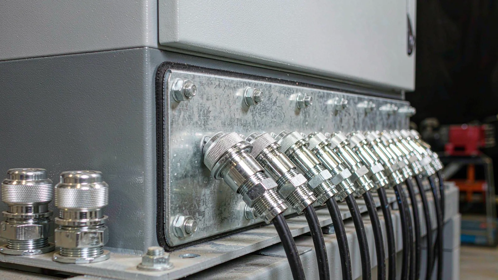 Galvanised gland plate bolted to the bottom flange of an electrical enclosure with a gasket line and a row of brass cable glands